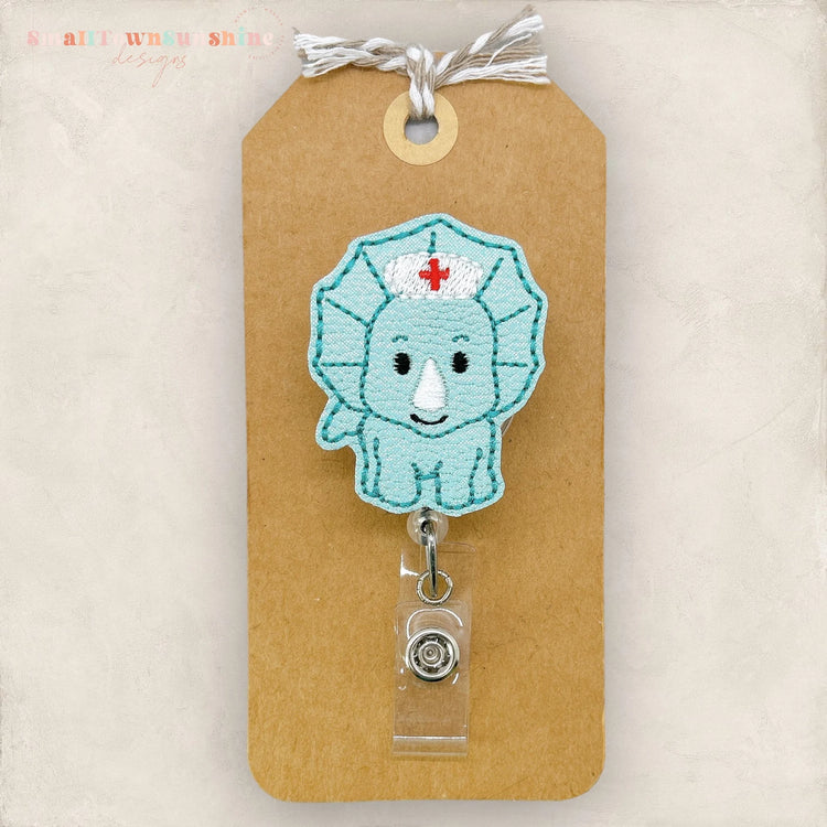 A handmade nurse badge with a blue triceratops design, attached to a brown paper tag.