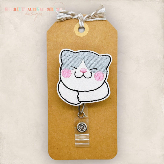 May include: A white and gray cat cartoon with pink cheeks, embroidered on a brown tag with a clear plastic retractable badge holder attached. The tag has the text 'Small Town Sue Shop Designs' at the top.